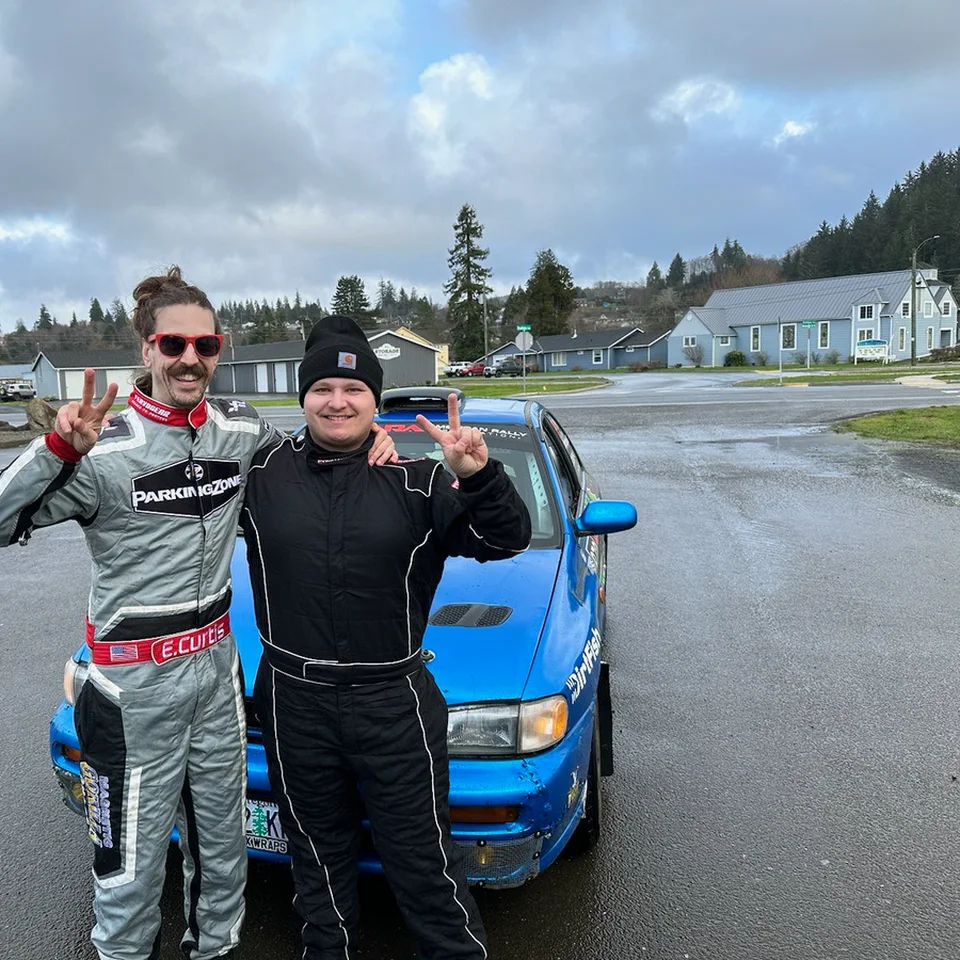 Two racers in FervoGear suits, one grey and one black, making peace signs beside their blue race car Two racers in FervoGear suits, one grey and one black, making peace signs beside their blue race car