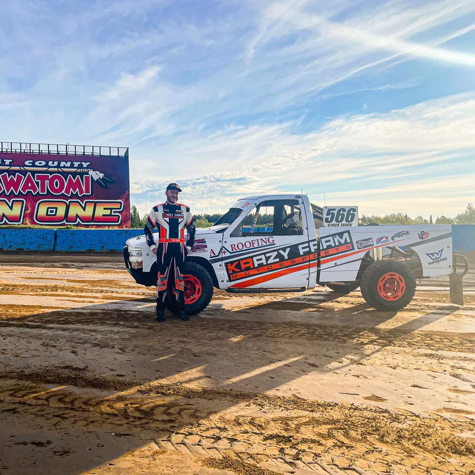 A driver in a red, white and black FervoGear suit beside his number 566 race truck in a dirt stadium A driver in a red, white and black FervoGear suit beside his number 566 race truck in a dirt stadium